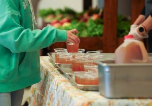 Delaware student in green sweatshirt picks up a sample of Delaware Grown apple cider from a table served by school nutrition staff, with local produce available in the background.