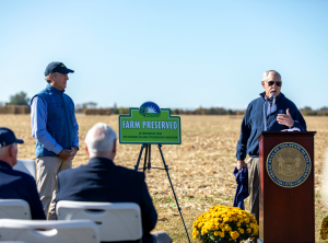 Governor Carney stands to the left of the new Aglands Preservation Farm sign as Delaware Secretary of Agriculture Michael T. Scuse speaks behind the podium with a farm field in the background and audience sitting in front.